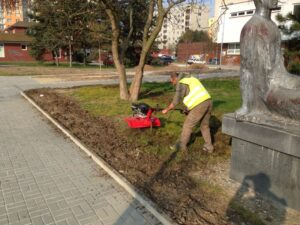 Osoba ve žluté bezpečnostní vestě obsluhuje červený rotační kypřič, aby zkultivovala půdu podél okraje chodníku v městském parku, v blízkosti stromu, velké sochy a moderního Bytového komplexu v pozadí.