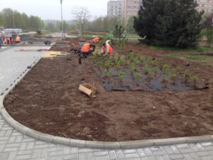 Dělníci v oranžových vestách sázejí malé keře a upravují terén podél dlážděné cesty u Bytového komplexu v městské oblasti, v pozadí jsou vidět stromy, budovy a zařízení.