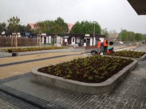 Několik pracovníků v oranžových vestách upravuje terén kolem vyvýšených zahradních záhonů podél dlážděného chodníku v městské části nedaleko Bytové komplexy, v pozadí stromy, budovy a přívěs.