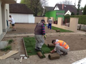 Několik lidí pokládá na dvoře před domem role drnového trávníku, připravuje a upravuje trávník. Další dělníci jsou poblíž a upravují terén. Jedná se o předměstskou čtvrť.