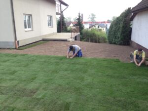 Dva lidé pokládají na zahradě drny a na holou půdu u domu pokládají zelené travnaté čtverce. Je vidět zahradnické nářadí a v pozadí jsou vidět domy a stromy.