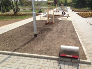 Upravená parková plocha v blízkosti bytové komplexy s čerstvou půdou, mladými stromky, lavičkami a kovovým válcem na dlážděném chodníku naznačuje nedávné zahradnické nebo terénní úpravy.