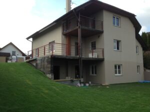 Dvoupatrový béžový dům se svažitým zeleným trávníkem, zastřešeným balkonem a vyvýšenou dřevěnou terasou s kovovým zábradlím. Ve spodním patře je několik oken a otevřené dveře.