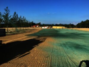 Staveniště se zelenými Hydroosev trávníky, roztroušenými stromy a několika budovami a zařízeními v pozadí pod jasnou modrou oblohou.