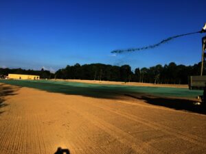 Velké otevřené pole je postřikováno zelenou látkou ze stroje vpravo, pravděpodobně pro Hydroosev trávníky nebo instalaci trávníku, se stromy a jasnou modrou oblohou v pozadí.