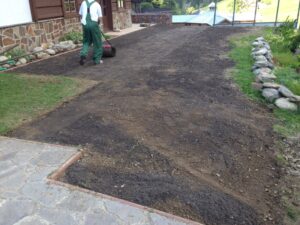 Osoba v zelené kombinéze zarovnává válečkem holou půdu na dvoře u domu a připravuje půdu pro výsadbu nebo terénní úpravy. Nedaleko jsou vidět kamenné obruby a cesta.