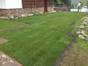 Čerstvě položený drn na dvoře vedle domu s kamennou a dřevěnou fasádou; na trávě leží zahradní hadice a v pozadí stojí člověk v blízkosti skal a hor.