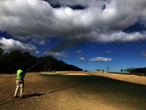 Osoba v neonové bezpečnostní vestě aplikuje Hydroosev trávníky a stříká tmavou látku na velkou, nově připravenou plochu pod částečně zataženou oblohou se stromy v pozadí.
