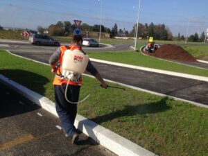 Pracovník v oranžové bezpečnostní vestě používá zádový postřikovač pro chemické ošetření trávníku poblíž silničního kruhového objezdu, v pozadí projíždějí auta a jiný pracovník obsluhuje stroje.