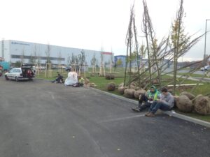 Několik lidí sedí na obrubníku parkoviště u stromků s kořeny připravenými k výsadbě. Poblíž stojí auto a vybavení a v pozadí je vidět průmyslová budova.