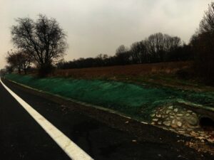 Vedle travnatého náspu s odvodňovacím potrubím, kde Hydroosev trávníky pomáhají udržovat bujný porost, vede venkovská cesta ohraničená bílou čarou. Stromy bez listí a zatažená obloha vytvářejí náladovou, pochmurnou atmosféru.